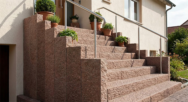 Robuste Außentreppe aus rötlichen Granit-Blockstufen mit Edelstahlgeländer am Hauseingang